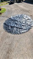 Black circular trampoline mat laid out flat on pavement under bright sunlight. The mat has visible attachment holes around the edge.
