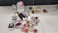 Overview of two Scentsy wax warmers and numerous small containers and clamshell packs of wax melts in assorted colors and scents arranged on table.