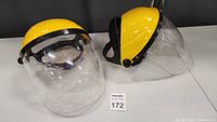 Photo of two new yellow headband face shields with clear visors and black straps, lying on a black surface with an auction lot number visible.