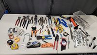 Full overview of tool lot showing pliers, wrenches, socket sets, clamps, tape measures and other assorted hand tools spread on a white table.