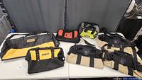 Seven tool bags displayed on a table: two DeWalt branded bags (yellow/black), one Black & Decker red/black bag, and four other bags in beige/black and yellow-green/black.