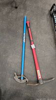 Blue pipe wrench and red pick mattock placed on concrete floor, both showing signs of usage and wear.