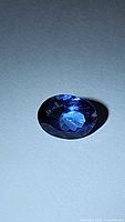 Close-up photo of dark blue oval cut tanzanite gemstone on a flat surface under light showing faceted cut and color.