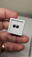Two round cut blue-teal-green sapphires in a white display box held by hand, showing the size and color of the gems clearly.