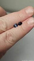 Pair of natural blue oval cut sapphires displayed on a finger, showing size scale and deep blue color.