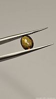 Close-up image of the oval cut sphene gemstone held by tweezers, showing its color and facet details from one angle.