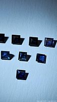 Eight natural blue sapphire princess cut gemstones arranged on a plain surface, showing size, color and clarity of each square-shaped stone.
