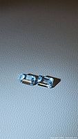 Close-up image of two natural aquamarine stones placed side by side, showing their emerald cut and clear blue color on a textured grey surface.