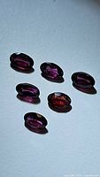 Six natural oval cut rhodolite garnet gemstones arranged on a flat surface showing purple pink color with facets visible.