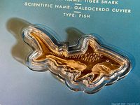 Close-up image showing the silver bar shaped like a hammerhead shark inside a custom clear capsule, against a booklet background with shark details.