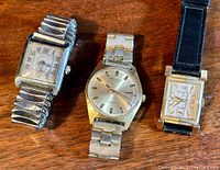 Three vintage watches on wooden surface, two Bulova rectangular watches and one Cardinal round watch with metal bracelet.