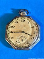 Top-down view of the pocket watch face, showing cream dial with Arabic numerals, the Lorie branding under 12 o'clock, and the distinctive small seconds subdial above 6 o'clock. Octagonal bezel with delicate floral engraving details visible.