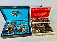Two vintage jewelry boxes open, showing assorted jewelry including rings, necklaces, bracelets, earrings, watches, and brooches