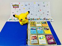 Blue binder album open to show vintage Pokemon trading cards in plastic sleeves placed on official Pokemon playmat with colorful Pokemon icons and characters.