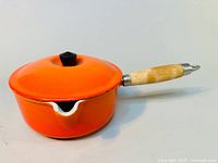 Side view of orange enamelled Le Creuset cast iron saucepan with wooden handle and lid, showing light chipping on the edge.