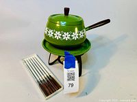 Photo of green aluminum fondue pot with white floral design, black wooden handle and knob, sitting on green drip plate and black tripod stand, alongside boxed new fondue forks with wooden handles.
