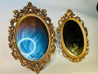 Two oval gold gilded framed mirrors hung on a wall, showing ornate decorative floral and scrollwork frames, vintage style.