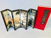 Full view of the six-panel miniature Chinese screen standing open, showing double-sided crane and nature artwork within black and gold decorative frames, next to the red box with gold and black labeling.