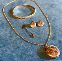 Full view of the jewelry lot laid on blue fabric: pendant locket on chain, bangle bracelet, pair of clip-on earrings, and stick pin.
