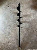 Full length top-down view of a black metal earth auger drill bit lying on tiled floor.