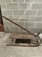Side view of antique wooden sled showing wood slat bed, metal frame, and long wooden handle leaning upwards