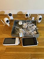 Lot of 4 white baby monitor cameras and 2 screens on wooden floor with a paper tray holding cords and accessories.