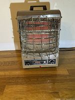 Front view of the compact Markel electric heater showing the protective metal grill, glowing red heating element, control knobs, and carrying handle on top.
