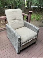 Outdoor rattan-style reclining chair with tan cushioned seat and back, shown on wooden deck in wooded outdoor area.