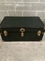 Closed view of rectangular green and black metal and wood trunk with metal locks and handles on concrete floor in front of cinder block wall.