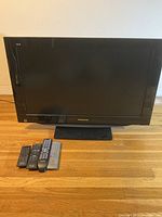 Full TV front view with the Panasonic LCD screen and the four remotes placed in front on wooden floor.