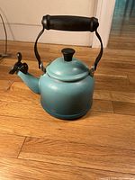 Blue enameled iron tea pot with black knob lid and handle, showing wear and discoloration on a wooden floor.