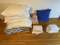 Full view of bed pillows stacked with couch pillows and folded pillowcases arranged beside on wooden floor.