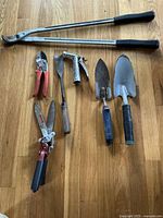 Photo showing the full set of 7 garden hand tools arranged on wooden floor. Tools include two hand trowels, pruning shears, lopper pruners, garden rake, spray nozzle, and hoe. Visible wear and rust on metal parts.