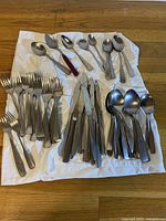Full collection of silverware including forks, knives, spoons laid out on a white cloth on a wooden table showing assorted styles and handle types.