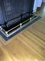 Side view of the traditional brass fireplace fender installed in front of a black fireplace hearth on a hardwood floor.