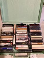 Full overhead view of mint green carrying case filled with multiple stacked cassette tapes, showing many different titles and artists.