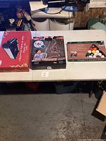 Three boxed games on a white table: a red box game, tabletop air hockey game, and a golfer's putter pool game.