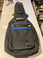 Black Gonher guitar backpack case with a blue stripe, front zippered compartments, and padded handle on table.