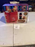 Front side of the boxed Nostalgia Electrics hot dog cooker with Coca-Cola design, displaying product image and branding.