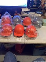 Nine new hard hats mostly in orange and some black, some wrapped in plastic, arranged on a table.
