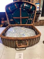 Wicker picnic basket open showing floral blanket and inside lid with green tableware attached.