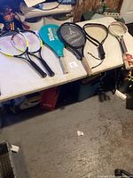 Five rackets placed on a table including two Wilson Matchpoint tennis rackets, one Wilson pickleball paddle, a black tennis racket with wooden handle, and a vintage wooden lawn tennis racket.