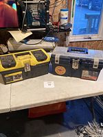 Two toolboxes on table: Stanley yellow/black toolbox on left and Homak black toolbox with clear top on right.