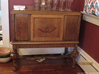 Front view of antique wooden buffet showing central carved door, side panels, metal handle, and carved apron with turned legs.