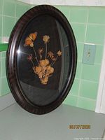Photo of antique oval wooden frame with dried flowers, showing front view with convex glass and dark wood finish.