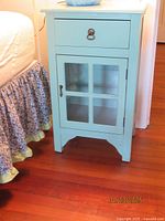 Front view of blue wooden accent cabinet showing one drawer with metal ring pull and one door with paned glass inserts beside a bed with floral bed skirt.