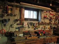 Wide view of the work bench, pegboard with hanging tools and dispensing items, shelves with toolboxes and containers below the bench.