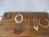 Various costume jewelry necklaces displayed on wooden surface including silver and gold chains, chokers, beaded and chunky necklaces