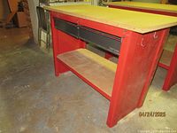 Full frontal view showing wooden top, red metal frame, two black drawers, and bottom shelf.