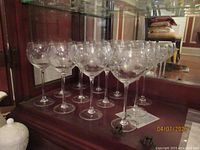 Twelve clear crystal stem wine glasses with etched decorative designs on the bowls arranged in a line on a wooden surface in front of a mirror.